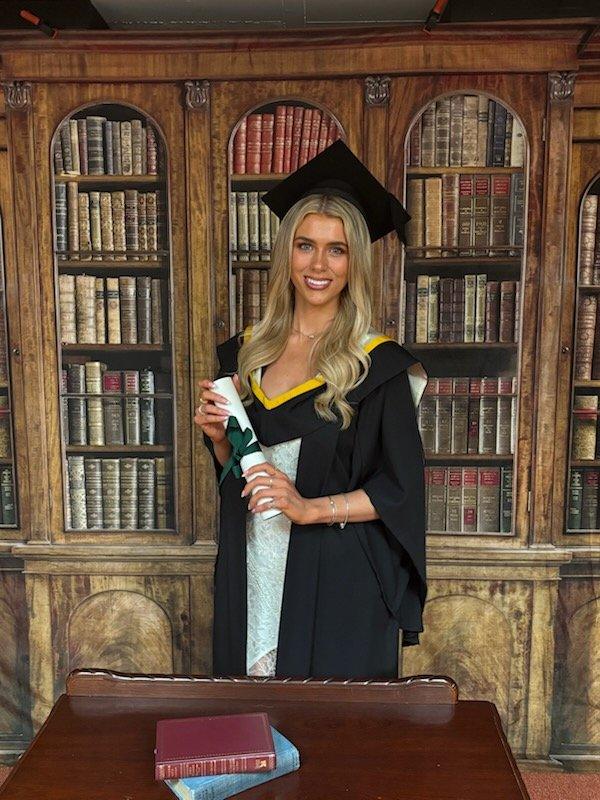 Aideen in her graduation cap and gown, holding her diploma in a library