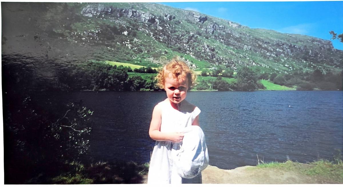 A young Aideen in a white dress beside an Irish lake, with green mountains beyond